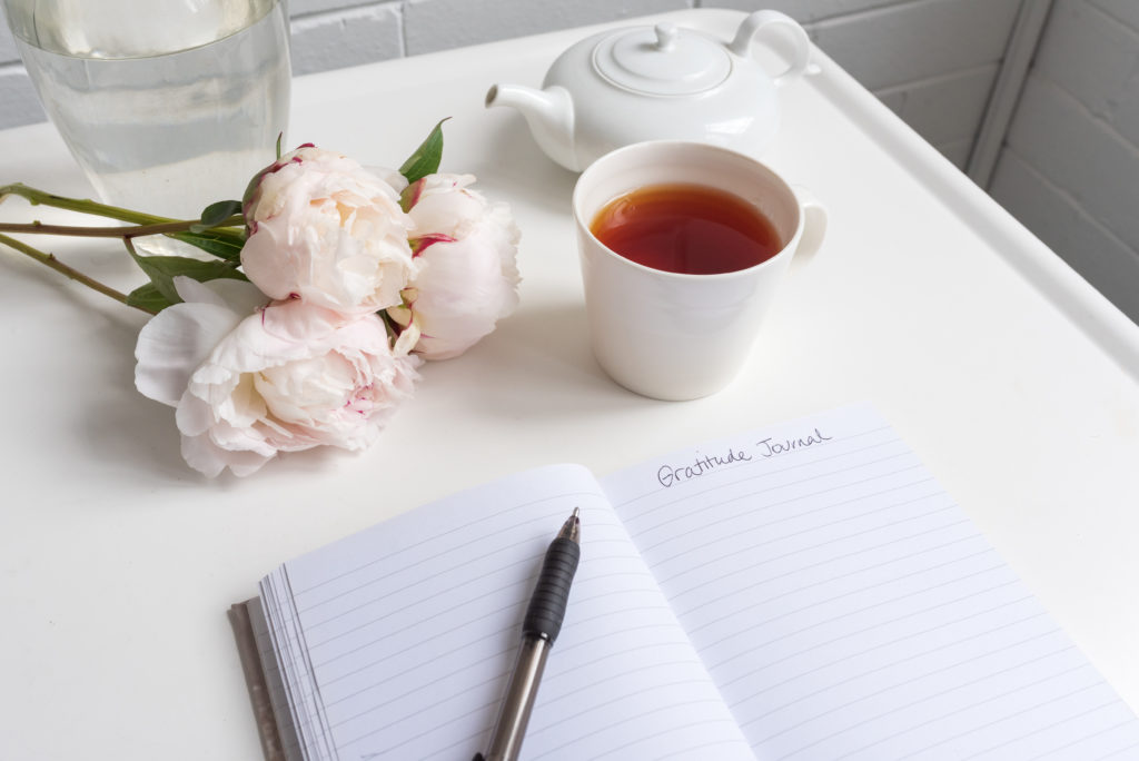 Closeup of gratitude journal on white table with peonies, cup of tea and teapot (selective focus)